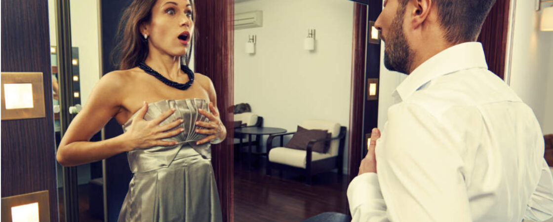 Eine überraschte Frau im Abendkleid steht vor einem Spiegel und unterhält sich mit einem Mann im Hemd.