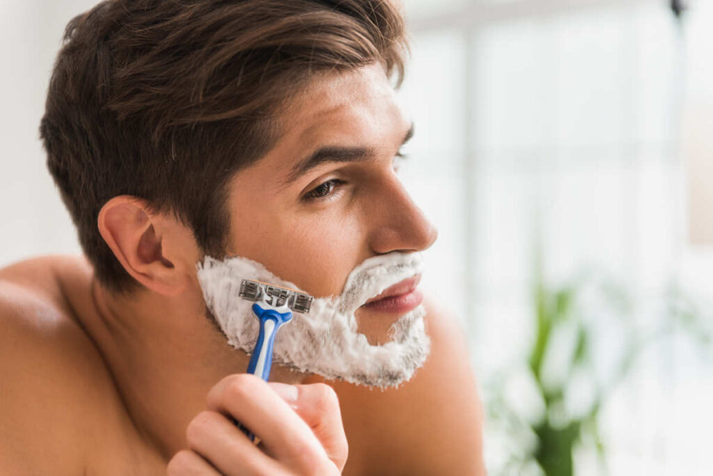 Ein junger Mann rasiert sich sorgfältig das Gesicht mit einem Rasierer und Schaum.