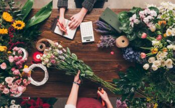 Zwei Personen arrangieren und planen Blumenarrangements auf einem Holztisch mit verschiedenen Blumen und Floristikwerkzeugen.