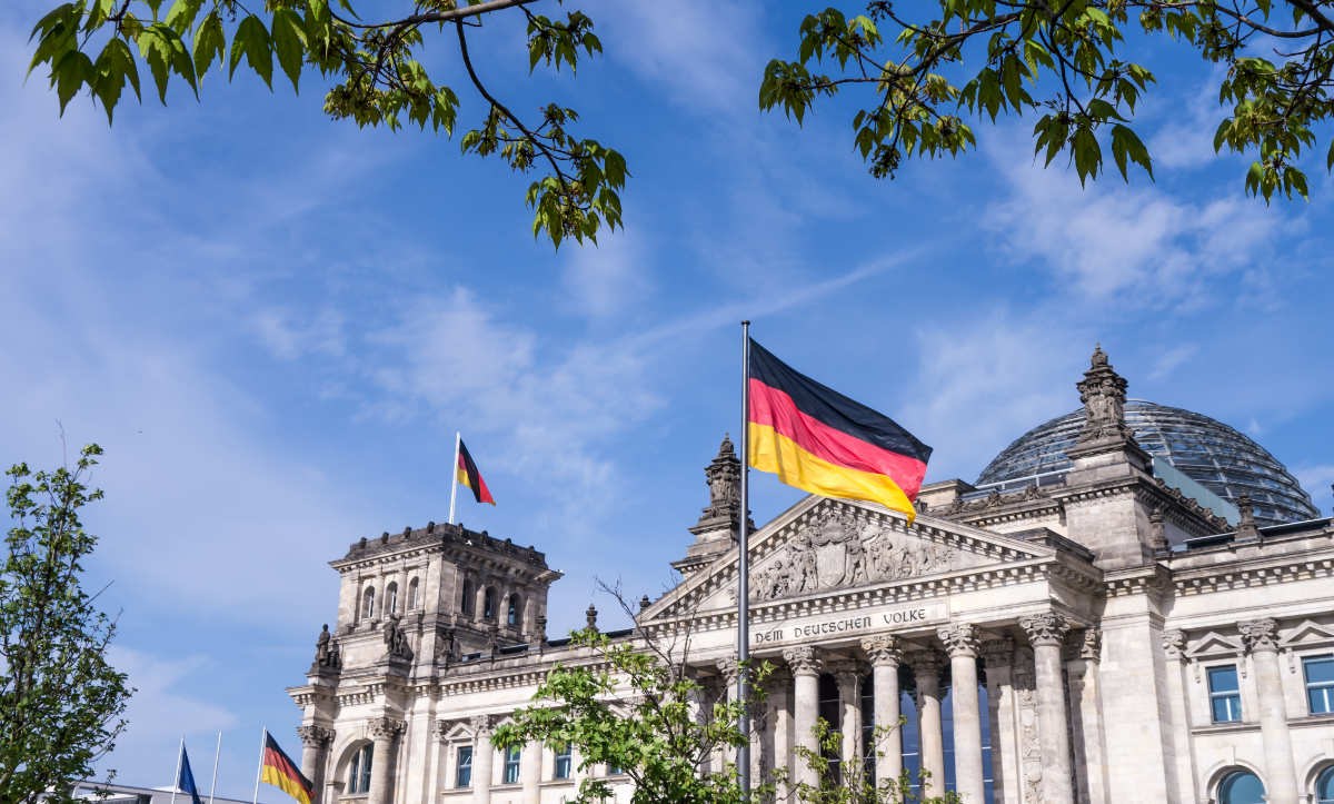 Das Bild zeigt das Reichstagsgebäude in Berlin mit wehenden deutschen Flaggen unter einem blauen Himmel mit einigen Wolken, eingerahmt von grünen Baumzweigen.